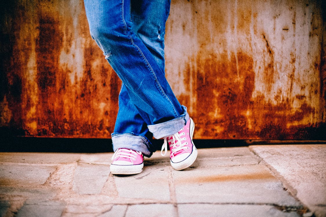 A woman stands against a rusty wall with jeans on and pink Converse tennis shoes with her legs crossed.