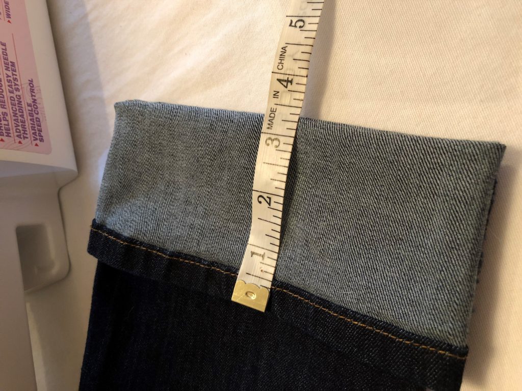 A pair of jeans lays on a sewing table with the bottom part of the pant leg folded up with a tape measure laying across it. How to hem jeans.
