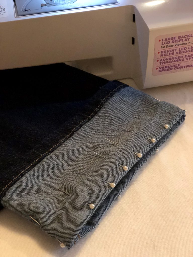 A pair of jeans lays on a sewing table with the bottom part of the pant leg folded up with pins spaced about one inch apart all the way around the pant leg to hold hem in place while sewing. How to hem jeans.