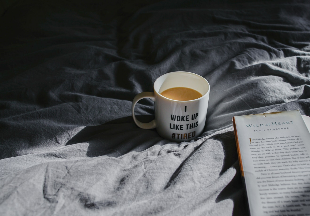 A cup of coffee that says I woke up this tired sits on a bed in the sheets next too an open book.