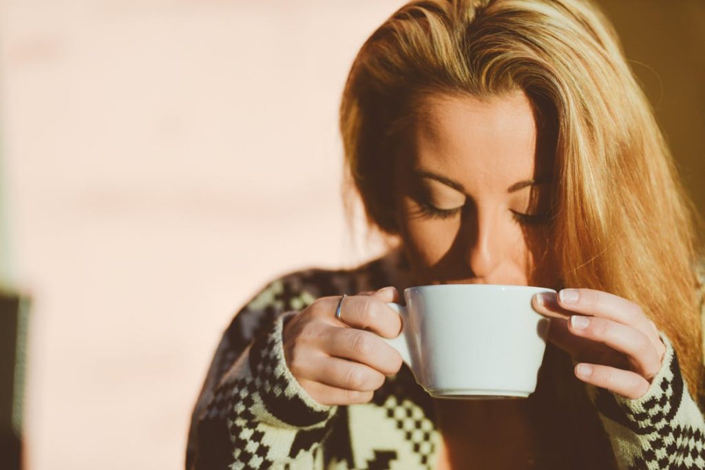 A blond woman with her hair down, looking down into the cup of coffee she is sipping, holding onto it with both hands.