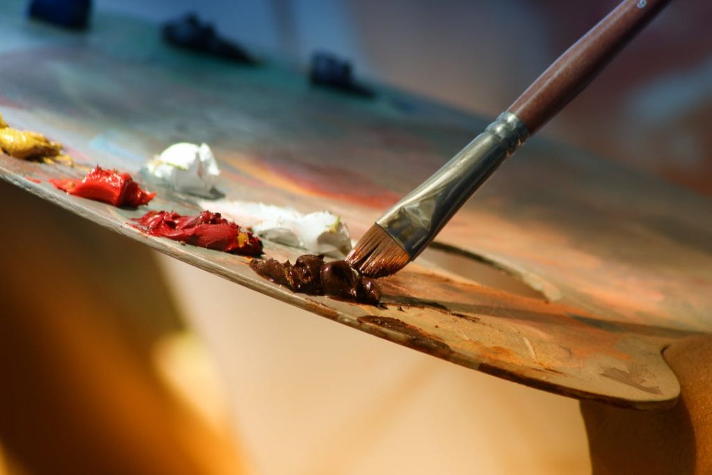 A paintbrush touches a wooden painter's palette , with white, yellow, red and orange paint on it.