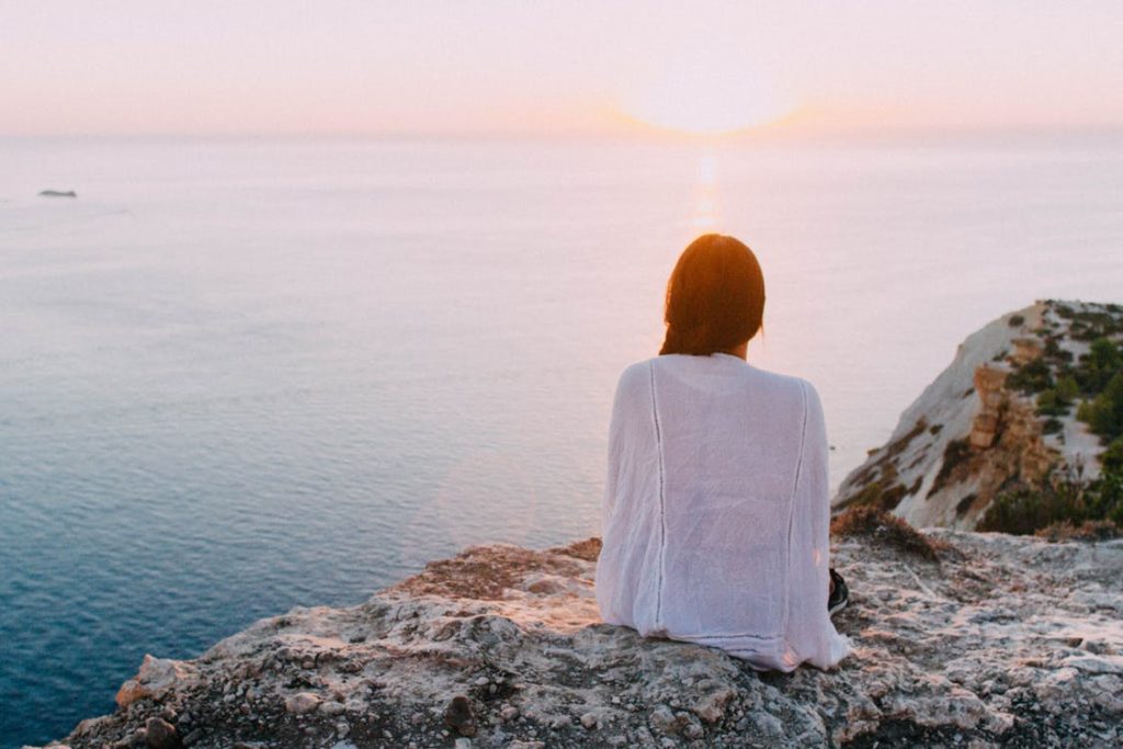 A young woman sits atop a cliff with a white shawl wrapped around her shoulders, looking out of over the water gazing into the sunset practicing mindfulness.