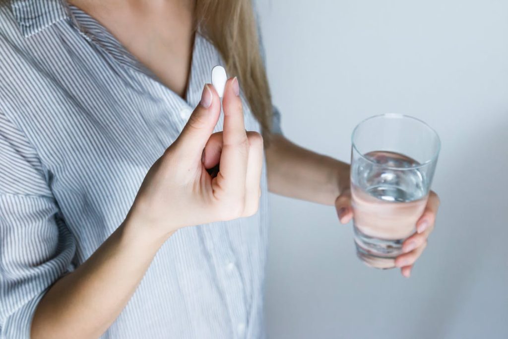 A woman in a gray shirt holding a glass of water in one hand and a pill in the other. 