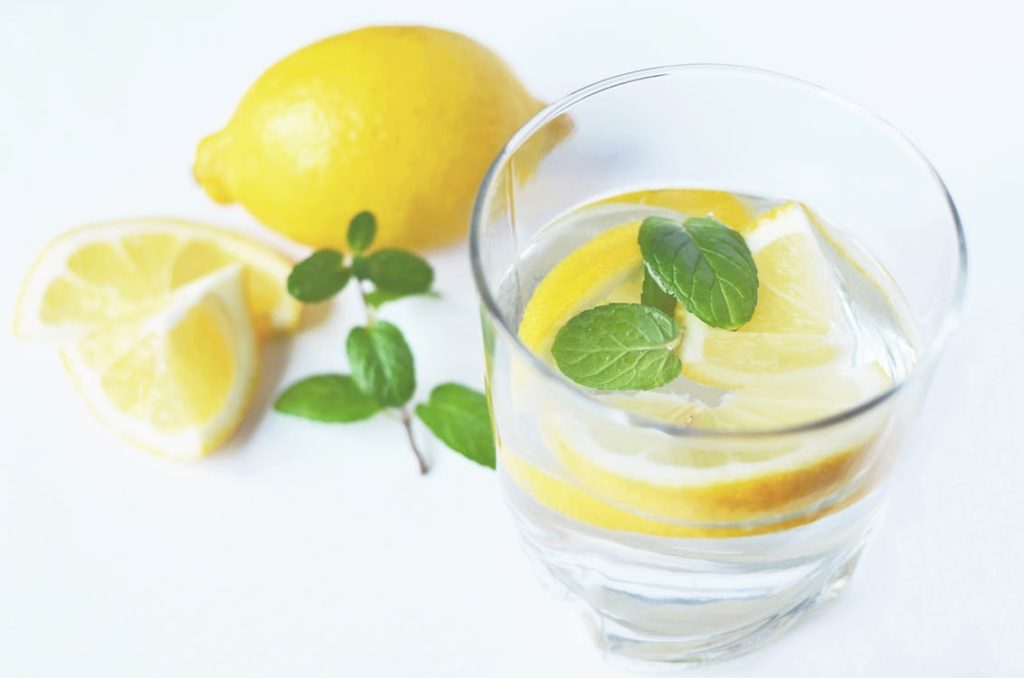A glass of water with cut up lemons and mint floating in it, set on a white table. 