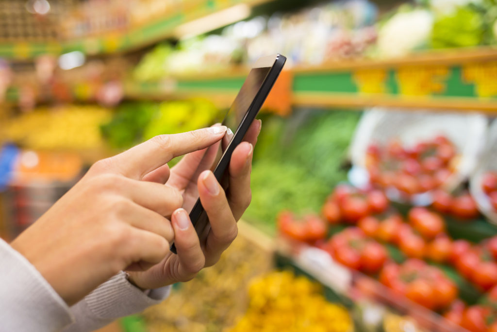 Woman using mobile phone while shopping in supermarket. Tips for frugal living and how to save money on groceries.