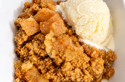 A serving of apple crisp and vanilla ice cream sit in a white bowl on a counter.