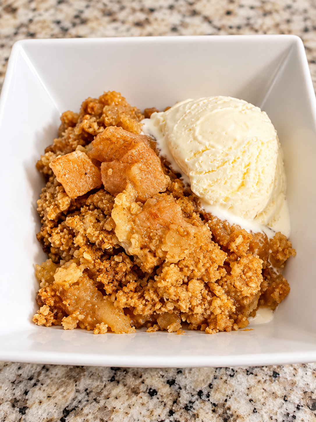 A serving of apple crisp and vanilla ice cream sit in a white bowl on a counter.