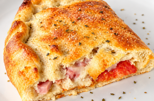 A baked ham and pepperoni calzone sits on top of a white plate.