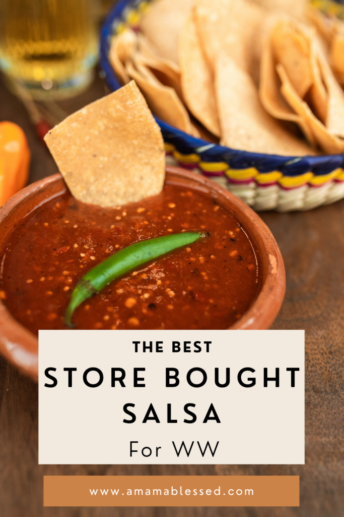 One tortilla chip stands upright in a small bowl of salsa, with one chili pepper on top. There is a bowl of tortilla chips in the background.