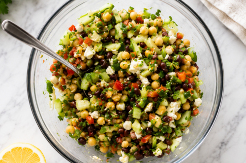 A mixed bean salad with beans, celery, peppers, chives, onions, cucumbers, and feta cheese sits in a bowl, on a counter next to a lemon wedge with a spoon in the bowl.