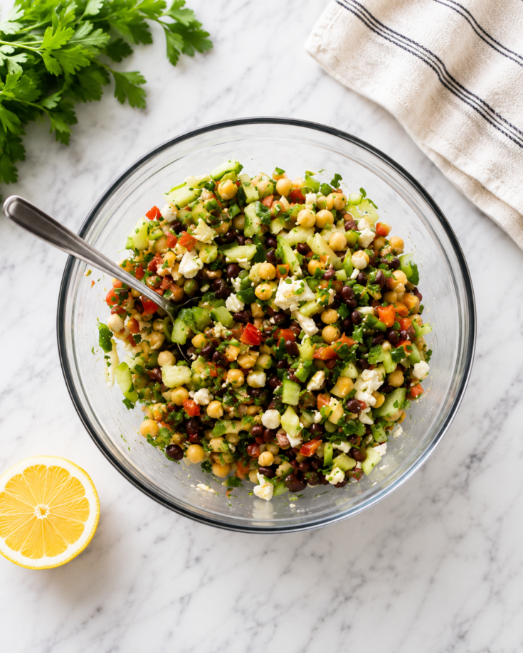 A mixed bean salad with beans, celery, peppers, chives, onions, cucumbers, and feta cheese sits in a bowl, on a counter next to a lemon wedge with a spoon in the bowl.