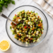 A mixed bean salad with beans, celery, peppers, chives, onions, cucumbers, and feta cheese sits in a bowl, on a counter next to a lemon wedge with a spoon in the bowl.