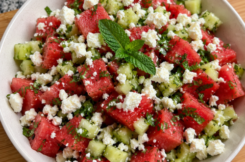 A watermelon salad with mint, cucumbers, and feta cheese is in a bowl sitting on a wooden cutting board on a counter.